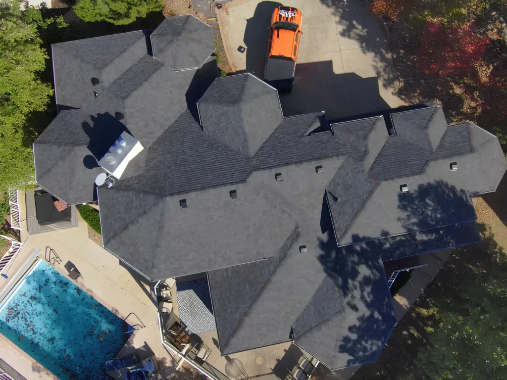 This image is showing an aerial view of a home with an intricate gray shingle roof, driveway, and backyard pool.
