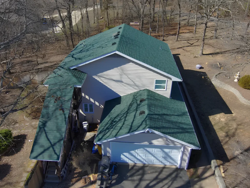 This image is showing an aerial view of a residential property featuring a green gabled roof and attached garage amid a wooded landscape.