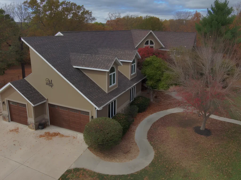 This image is showing a custom home in Lake Ozark with a newly installed dark gray architectural shingle roof, featuring multiple dormers, a two-car garage, and beautiful landscaping surrounded by fall foliage in the Ozarks region.