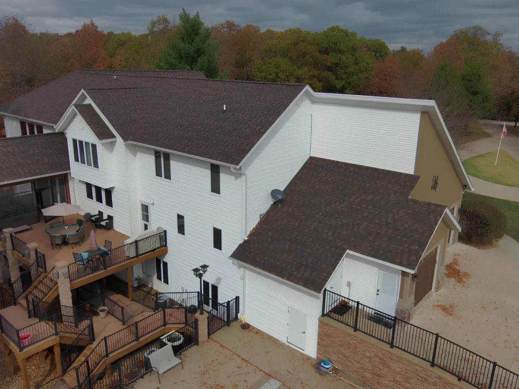 This image is showing a multi-level white home with brown shingle roof, custom deck installation, and multi-tiered outdoor living spaces surrounded by fall foliage in Lake Ozark.