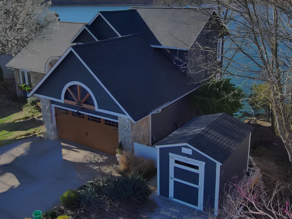 This image is showing a lakefront property with charcoal gray roof, stone accents, wooden garage door, and matching storage shed overlooking Lake of the Ozarks blue waters.