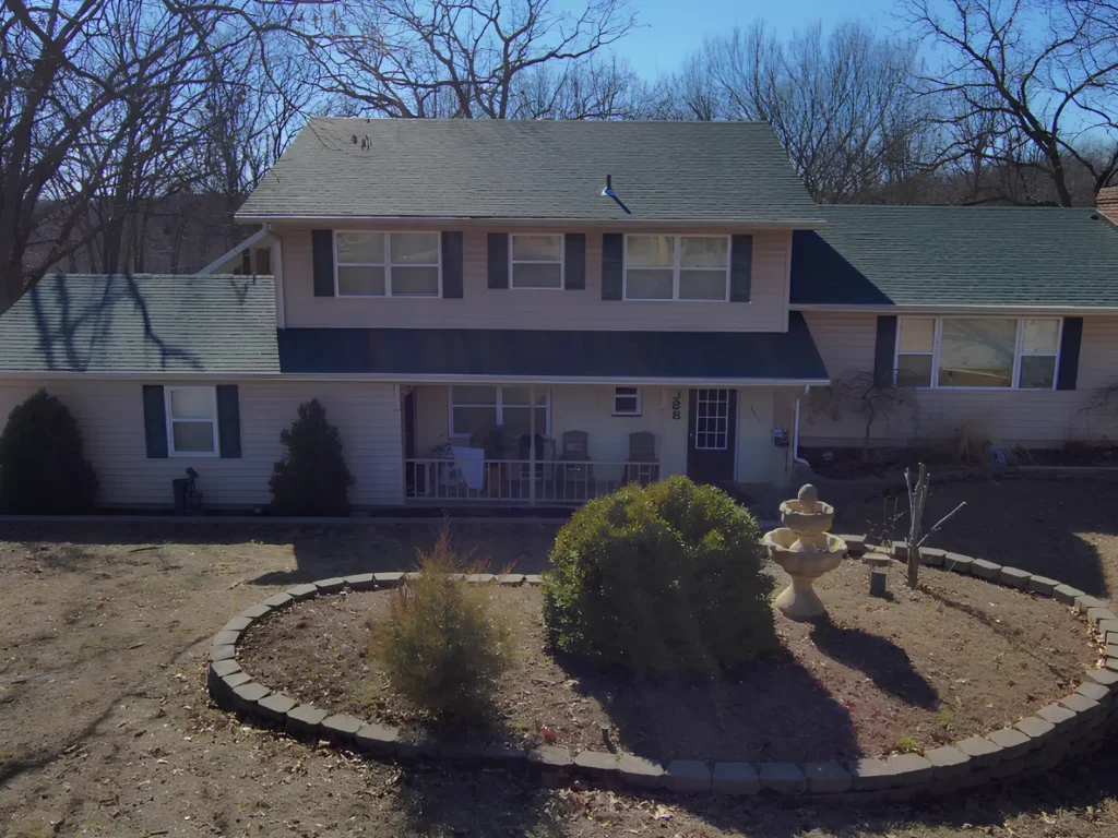 This image is showing a two-story residential home with new green roof installation, front porch, and circular landscaping feature in the Lake Ozark region.