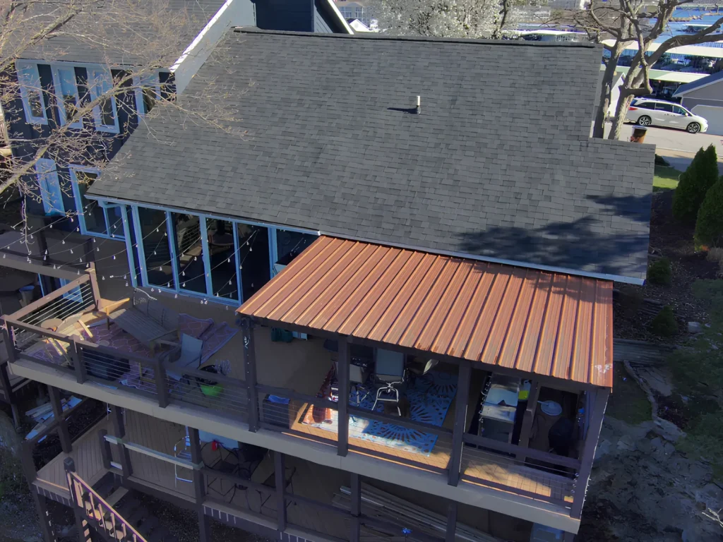 This image is showing a lakefront property with gray shingle roof and copper metal roof extension over a deck area, featuring modern architectural design elements common in Lake Ozark vacation homes.