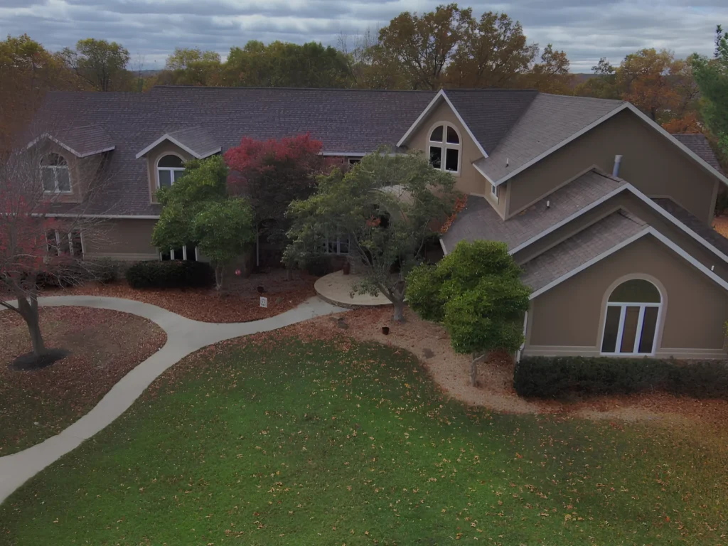 This image is showing an elegant custom home with architectural shingle roof, multiple dormers, and beautiful landscaping surrounded by fall foliage in the Lake of the Ozarks region.